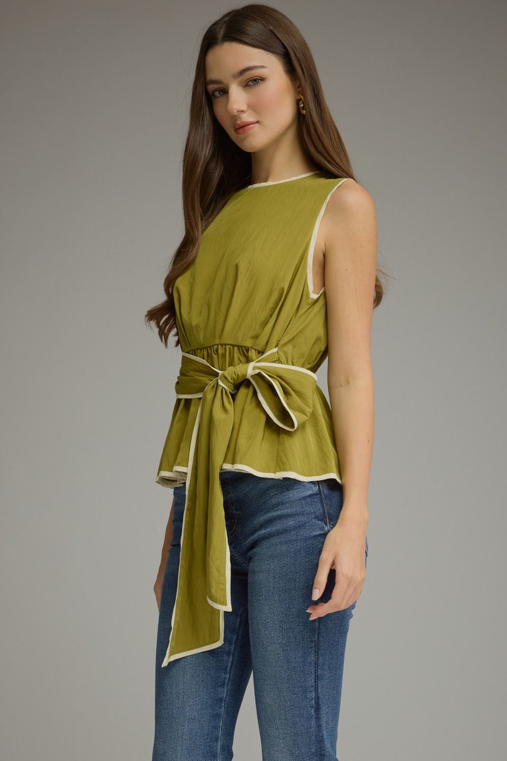 A woman modeling a sleeveless olive green top with cream trim and a matching tie belt, paired with blue jeans, against a solid gray studio background.