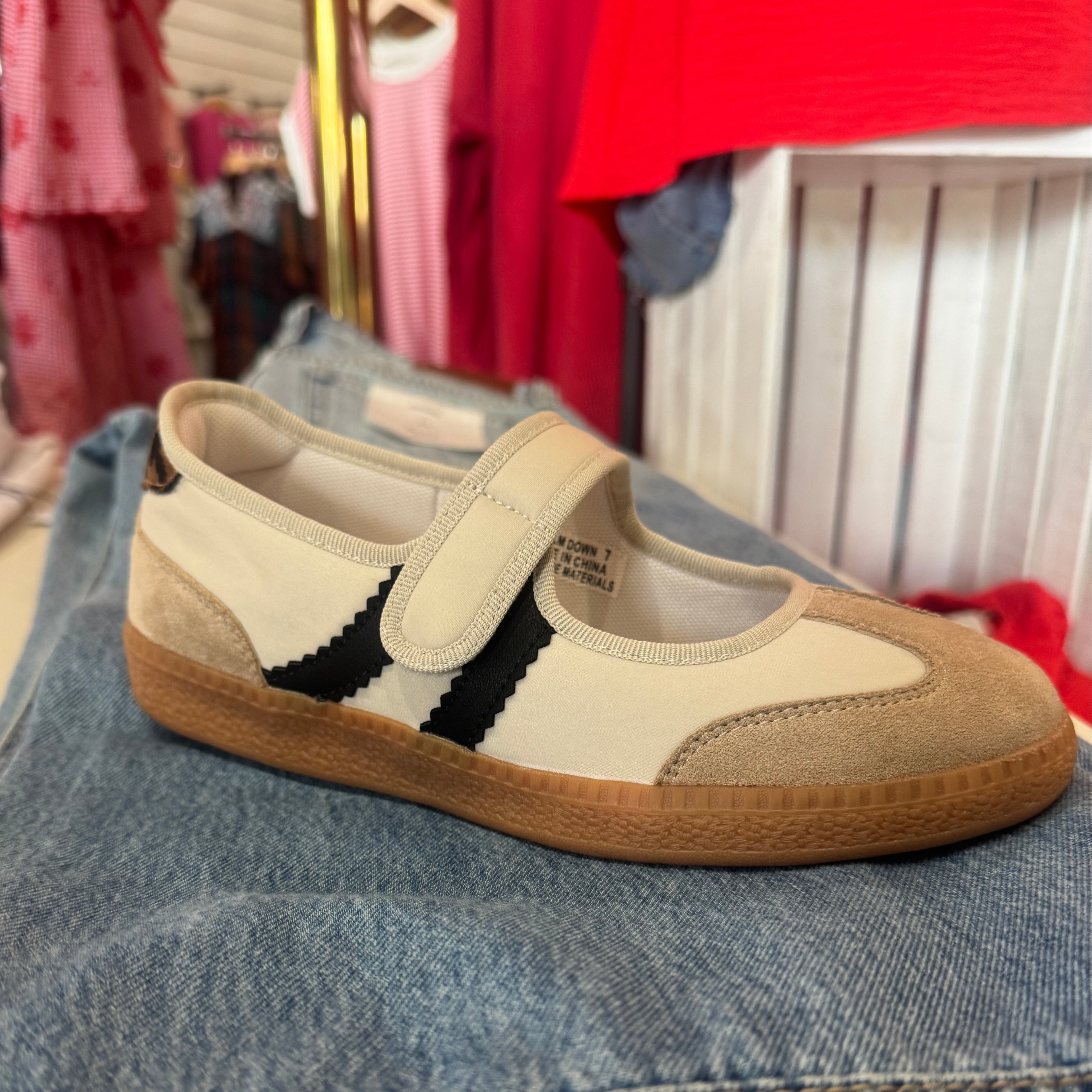 A single low-top shoe with a cream canvas upper, beige suede toe and heel panels, a black side stripe, and a brown gum sole, resting on a pair of blue jeans in a retail setting.