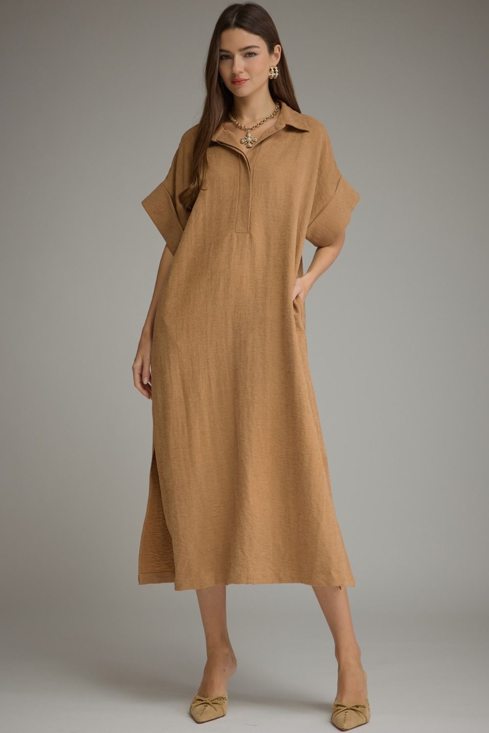 A woman modeling a loose, midi-length tan dress with a short zipper at the collar, short sleeves, and side pockets, photographed against a neutral gray studio background.