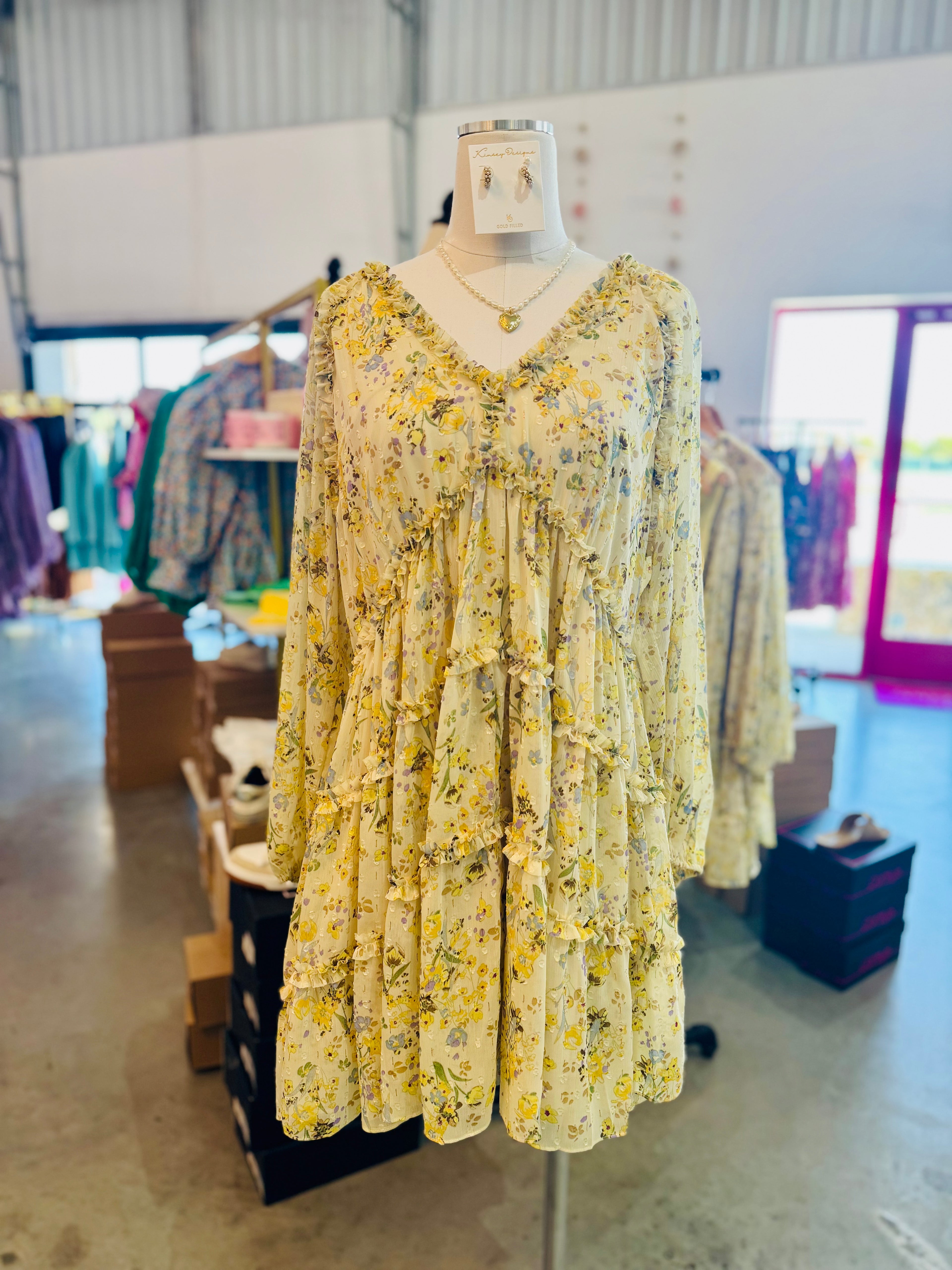 This image features the Carmina Tiered Dress, a charming and stylish garment displayed on a mannequin. The dress is adorned with a vibrant yellow floral pattern, adding a cheerful and lively touch.