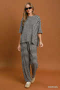 A woman modeling a charcoal and white striped lounge set consisting of an oversized short-sleeve top and matching wide-leg pants, paired with beige sneakers against a solid brown studio backdrop.