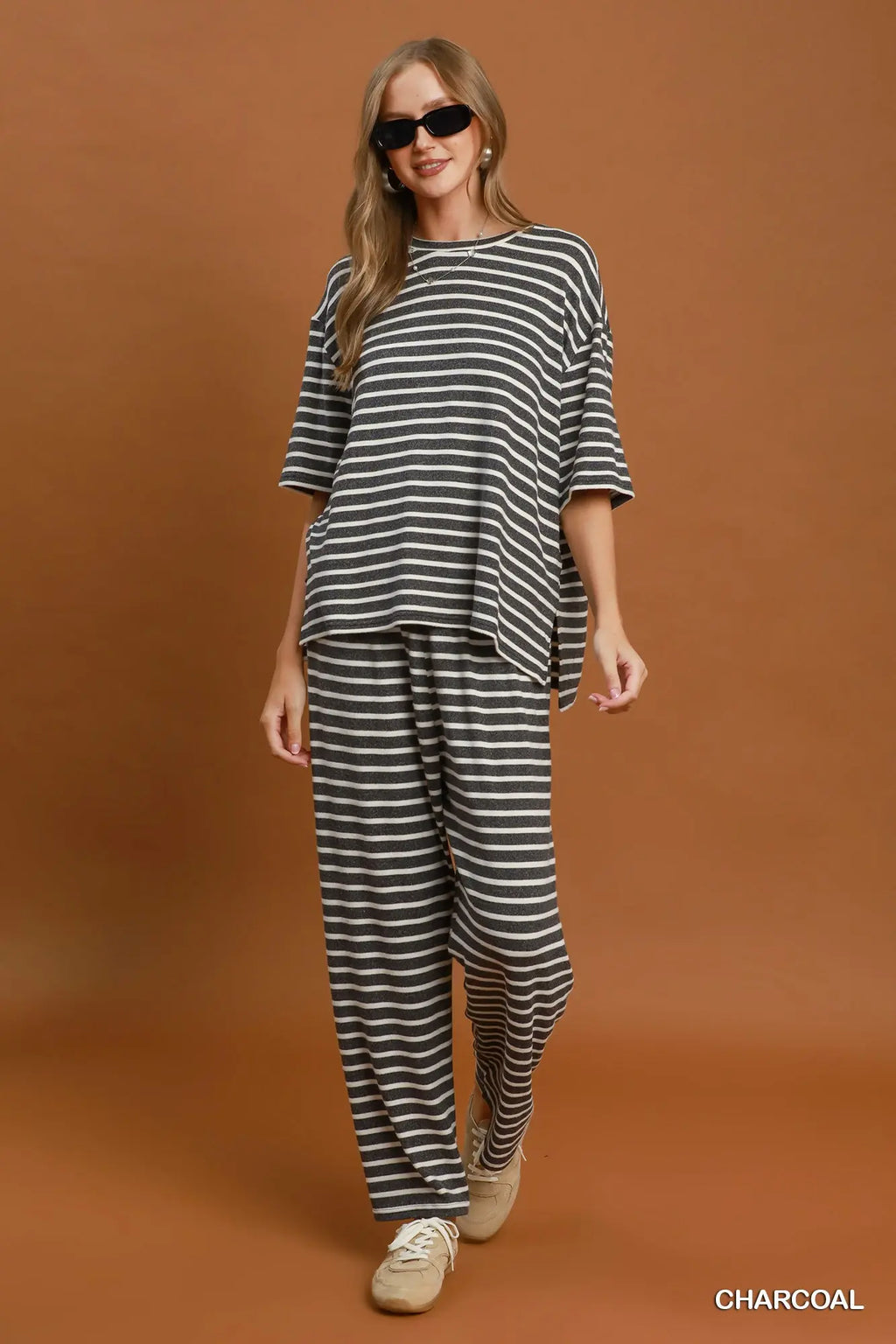 A woman modeling a charcoal and white striped lounge set consisting of an oversized short-sleeve top and matching wide-leg pants, paired with beige sneakers against a solid brown studio backdrop.