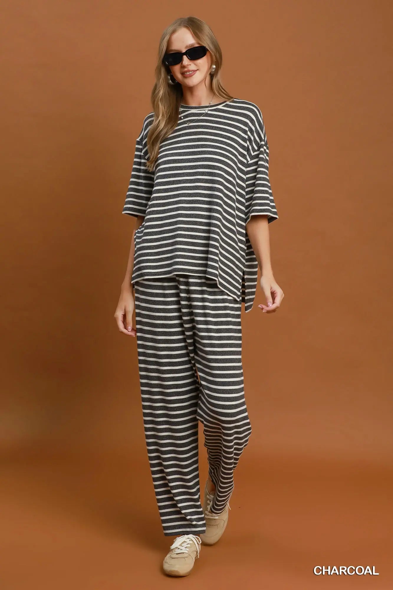 A woman modeling a charcoal and white striped lounge set consisting of an oversized short-sleeve top and matching wide-leg pants, paired with beige sneakers against a solid brown studio backdrop.