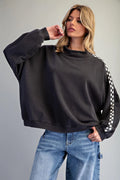 A woman modeling a dark charcoal gray oversized sweatshirt with a black and white checkered stripe detail on the sleeve, paired with blue jeans against a solid gray studio background.
