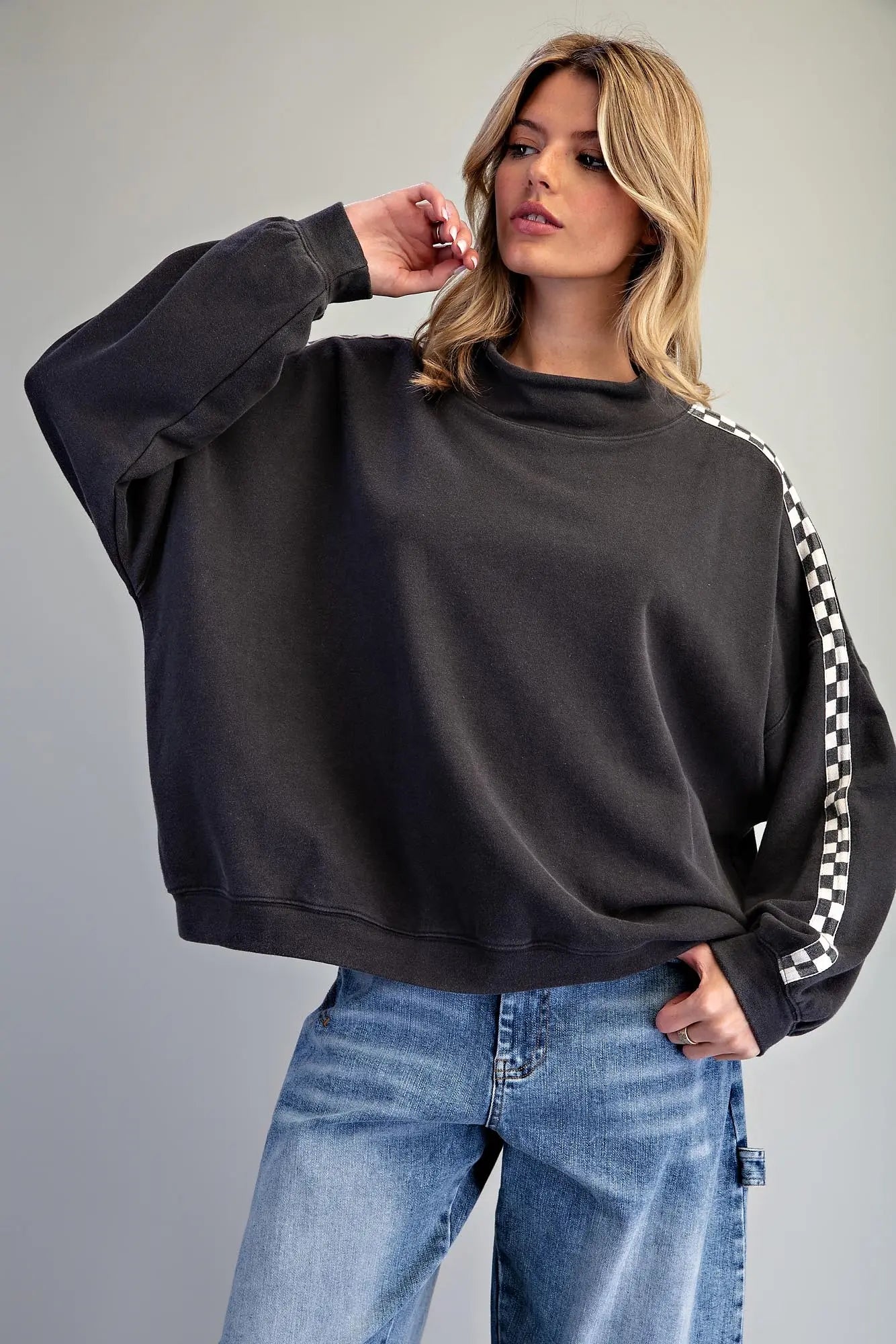 A woman modeling a dark charcoal gray oversized sweatshirt with a black and white checkered stripe detail on the sleeve, paired with blue jeans against a solid gray studio background.