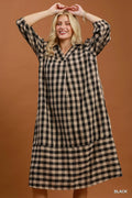 A woman modeling a black and beige gingham plaid long-sleeve shift dress against a solid warm brown studio background.