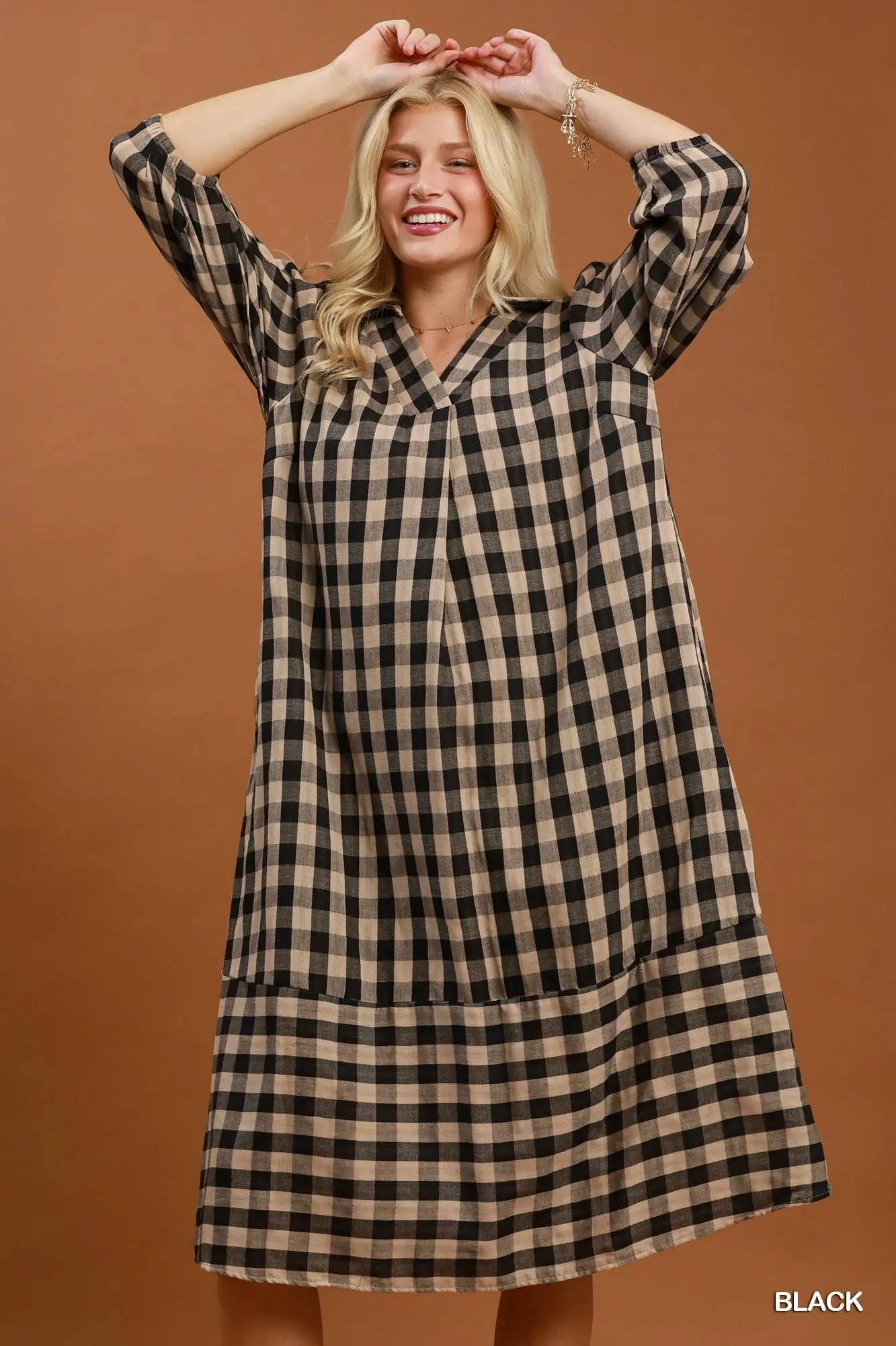 A woman modeling a black and beige gingham plaid long-sleeve shift dress against a solid warm brown studio background.