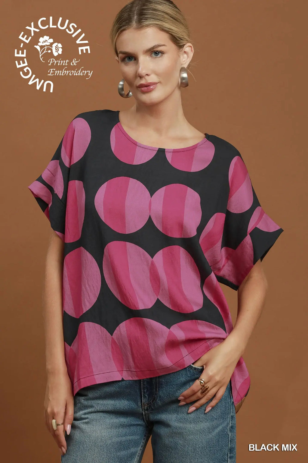 A woman modeling a short-sleeve top with a black base and large overlapping pink circles, paired with blue jeans, against a solid brown studio background. A logo in the top left reads 'UNLEASHED Print & Embroidery', and 'BLACK MIX' appears in the bottom right corner.