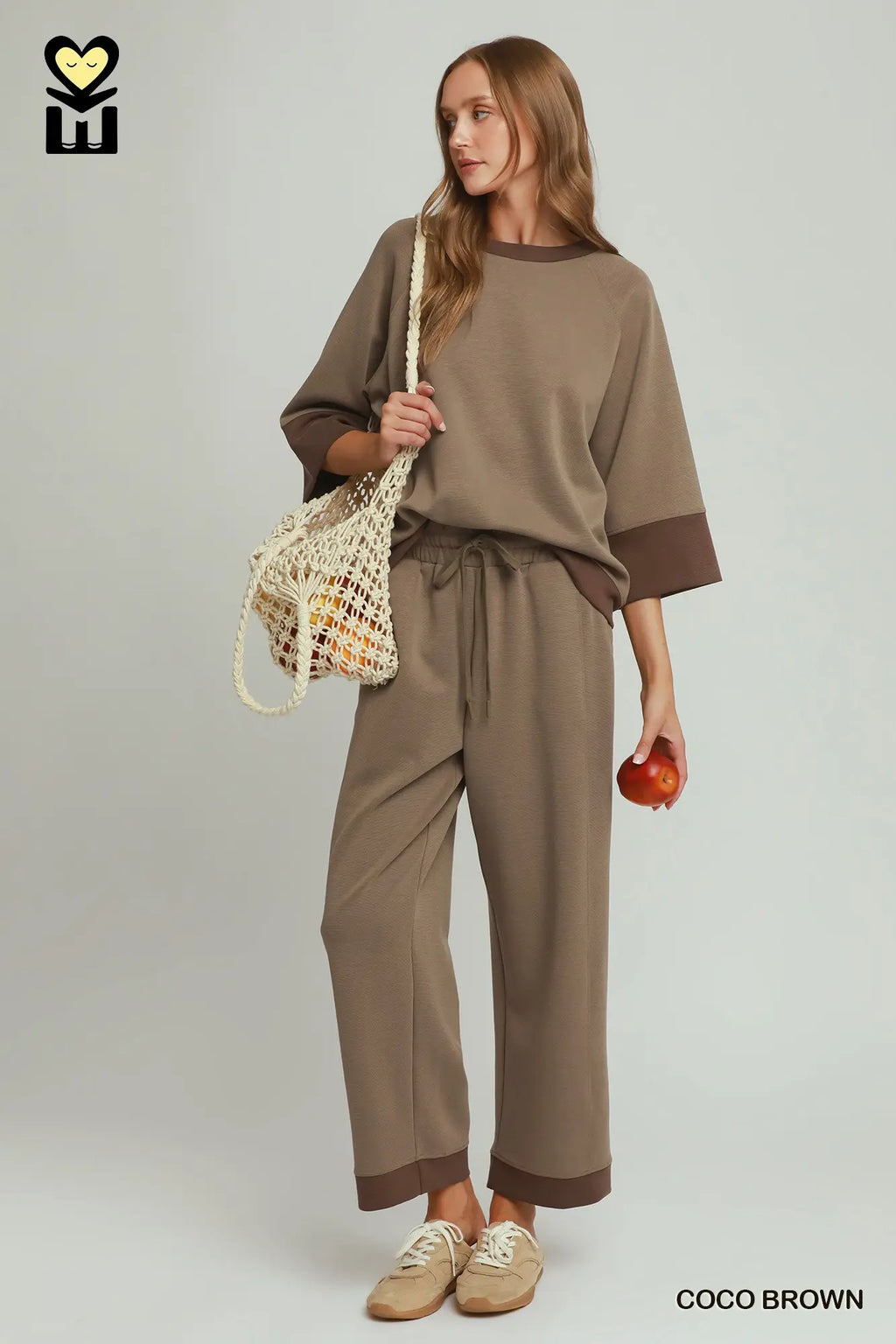 Woman wearing a brown outfit holding a bag and an apple against a plain background