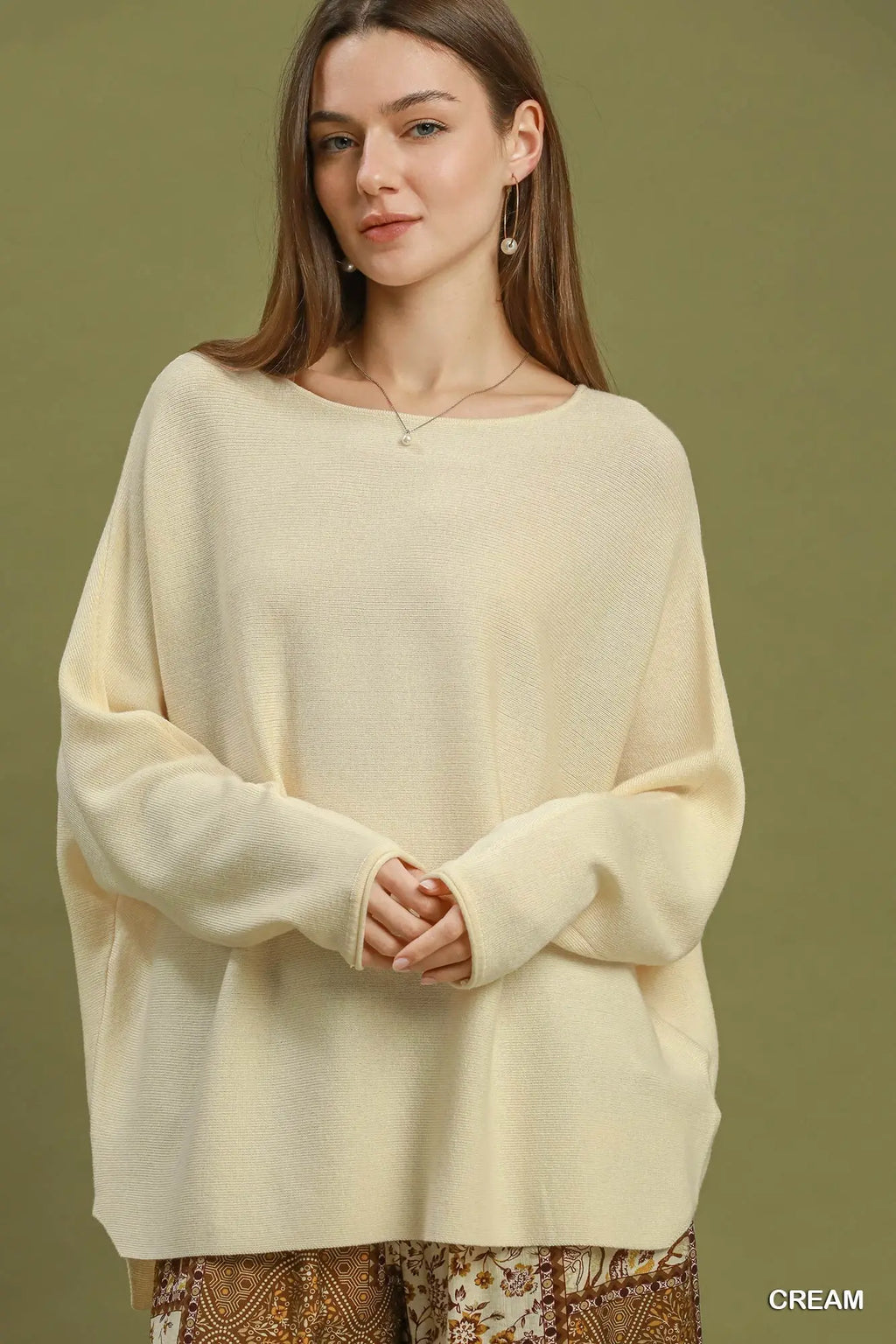 A woman wearing a cream-colored, oversized knit sweater with a wide boat neckline and long sleeves, posed against a solid olive green background.