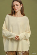 A woman wearing a cream-colored, oversized knit sweater with a wide boat neckline and long sleeves, posed against a solid olive green background.