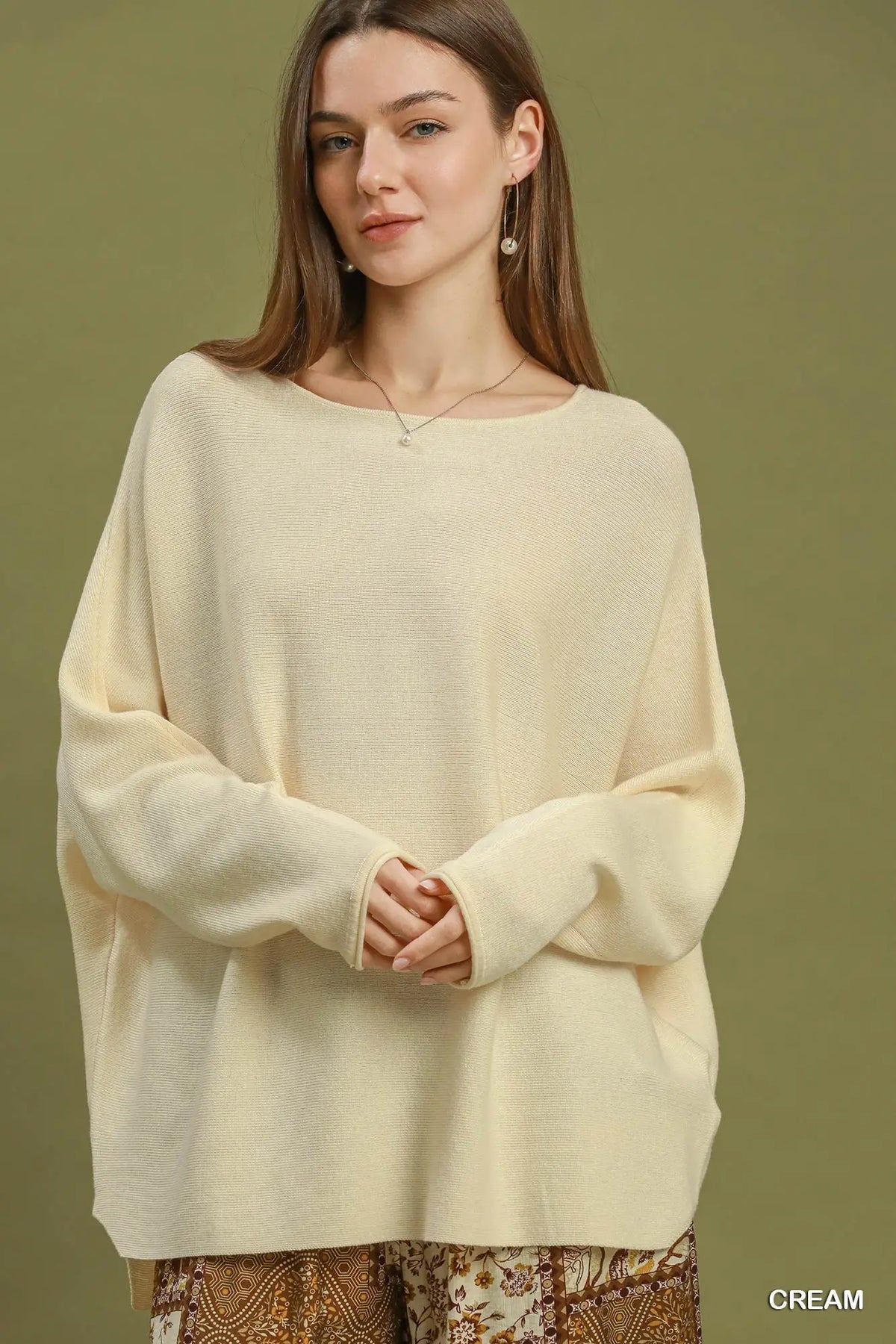 A woman wearing a cream-colored, oversized knit sweater with a wide boat neckline and long sleeves, posed against a solid olive green background.
