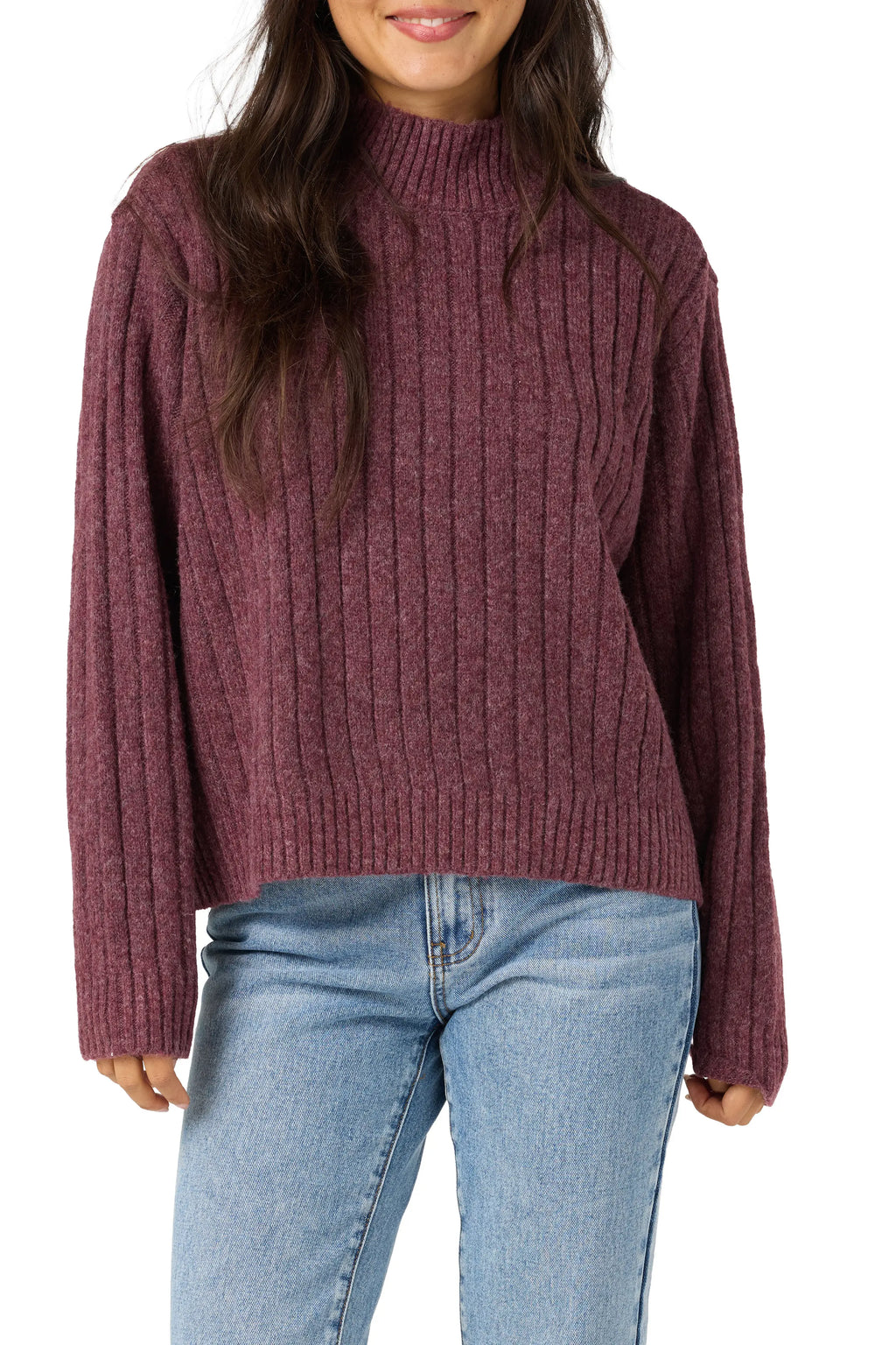 A woman wearing a ribbed knit mock neck sweater in a heathered burgundy color, paired with light blue jeans, against a plain white background.