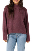 A woman wearing a ribbed knit mock neck sweater in a heathered burgundy color, paired with light blue jeans, against a plain white background.