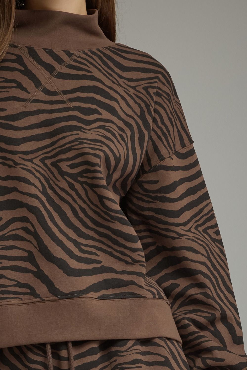 Close-up of a brown and black zebra-patterned sweatshirt with a mock neck and ribbed hem, worn by a model against a plain gray background.