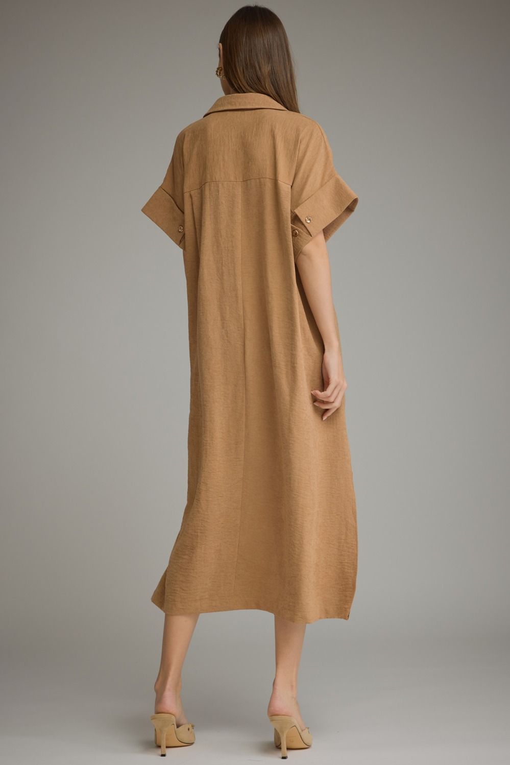 A woman viewed from behind wearing a loose, short-sleeve midi dress in camel brown, paired with beige heeled sandals, against a solid gray studio background.