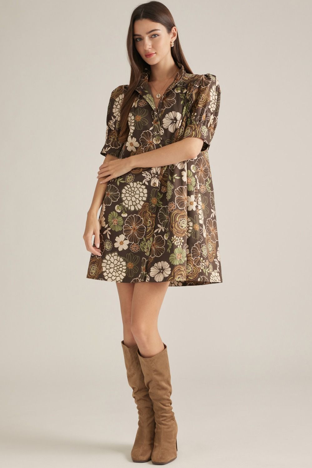 A woman modeling a short-sleeve mini dress with a brown, beige, and green floral pattern, paired with brown suede knee-high boots, against a solid light beige studio background.