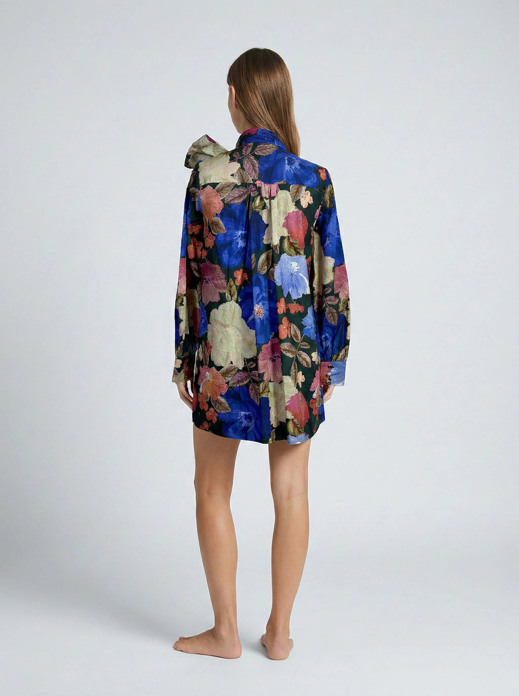 A woman stands barefoot against a light gray studio background, viewed from the back, wearing a long-sleeve floral robe or shirt in rich blue, pink, cream, and olive tones.
