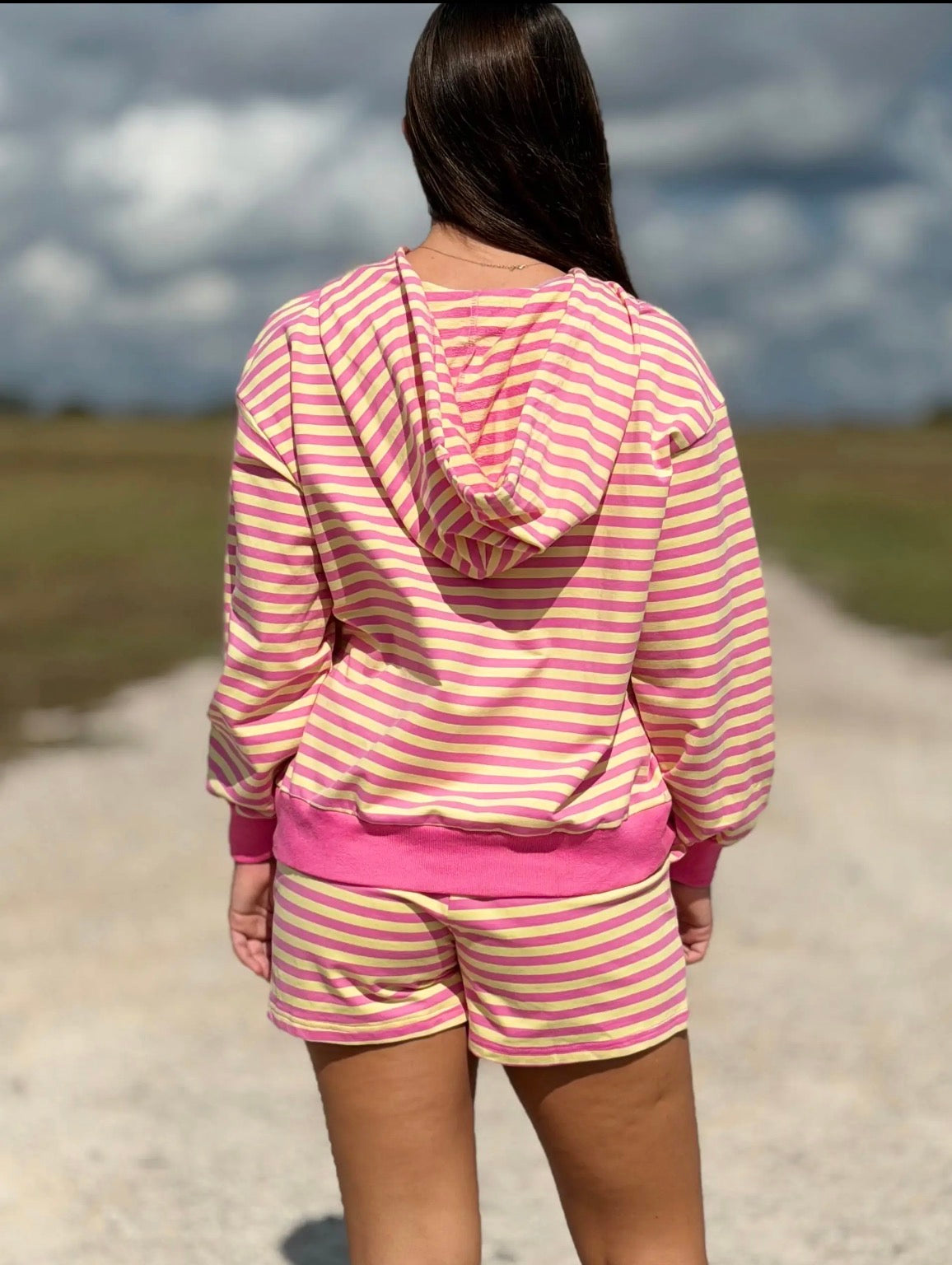 A person viewed from behind wearing a pink and white striped hooded sweatshirt and matching shorts, standing on a gravel path outdoors with a cloudy sky in the background.