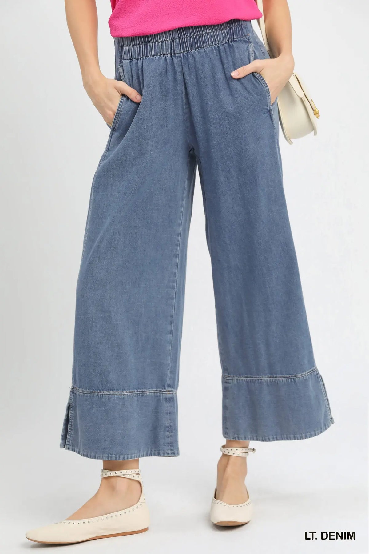 A model wearing light blue wide-leg denim pants with a gathered elastic waistband and rolled cuffs, paired with a pink top and cream flat shoes against a light gray studio background.