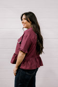 A woman with long dark hair is shown from the side/back wearing a short-sleeve ruffled blouse with thin vertical stripes in burgundy and white, paired with dark jeans, against a white paneled wall.