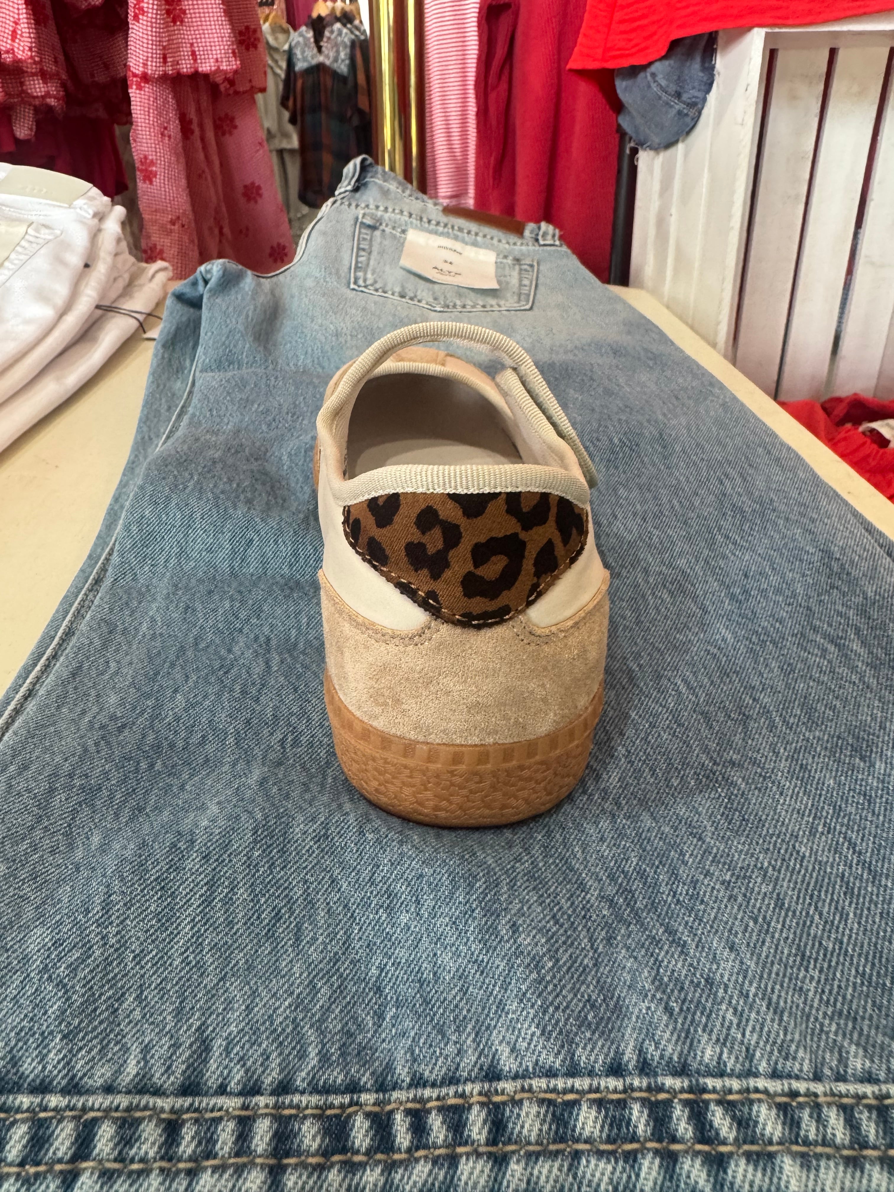 A single slip-on platform shoe with a beige suede upper, a leopard print heel accent, and a brown textured sole, displayed on top of a pair of blue jeans in a retail setting.