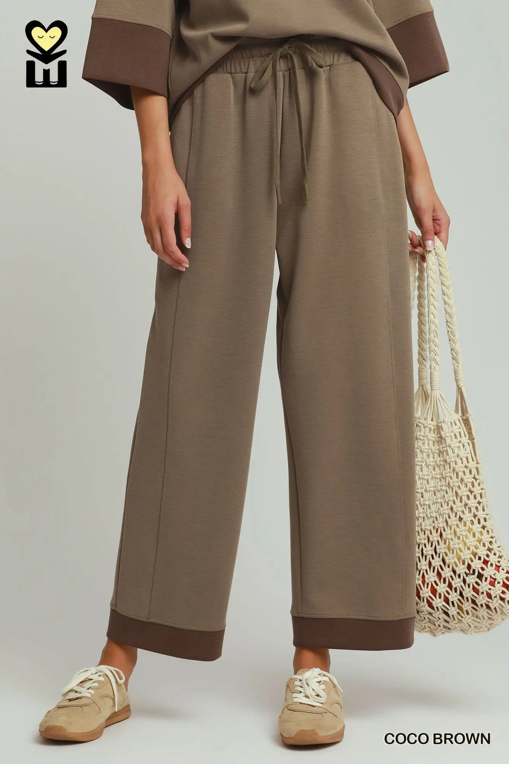 Person wearing a brown outfit with wide-leg pants holding a woven bag on a plain background