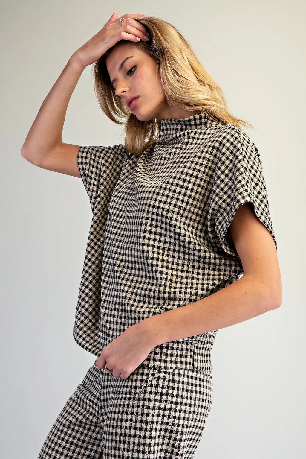 A woman modeling a black and beige gingham short-sleeve top and matching trousers against a plain light gray studio background.