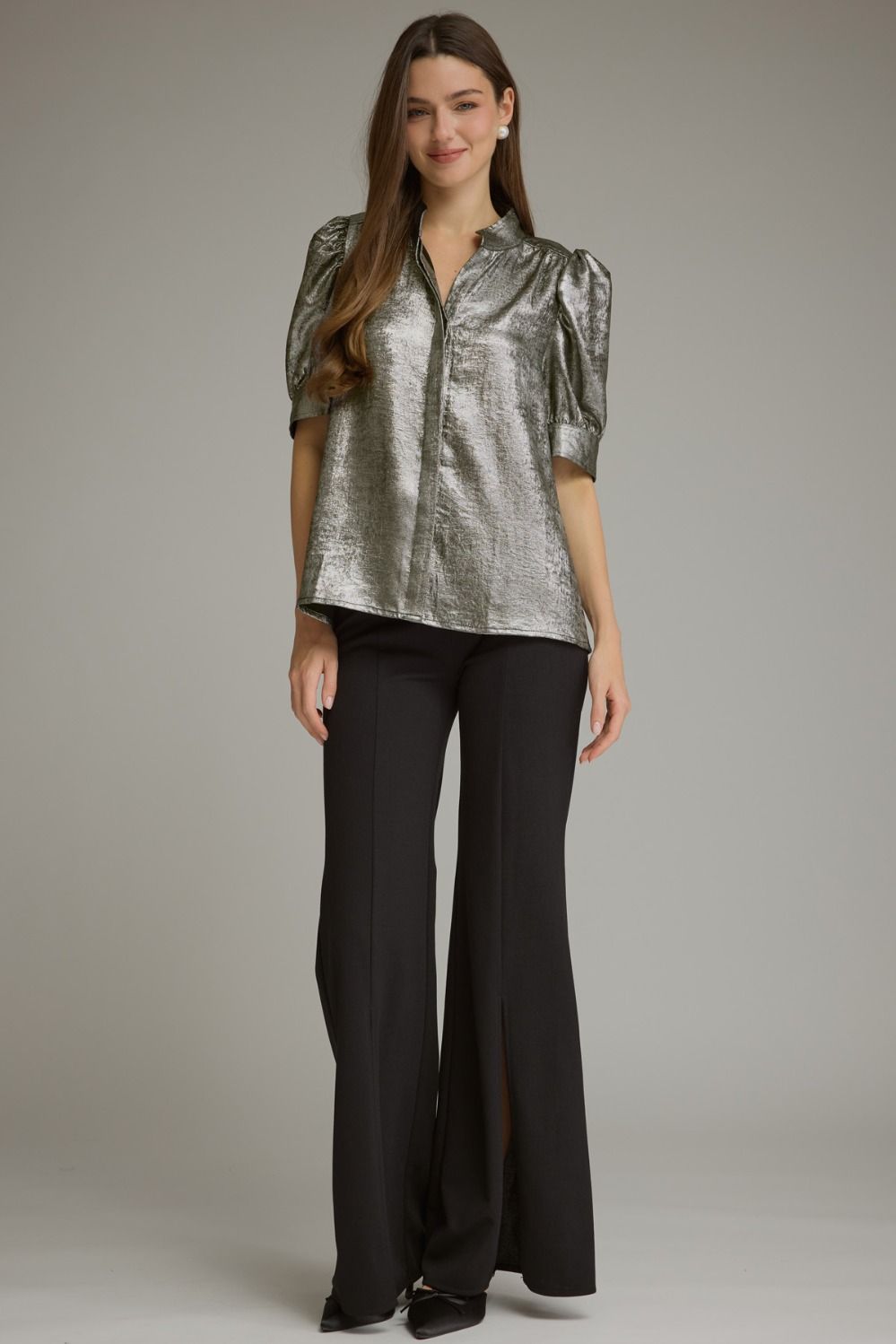 A woman modeling a metallic silver short-sleeve blouse with puffed sleeves and black wide-leg trousers, standing against a neutral gray studio background.