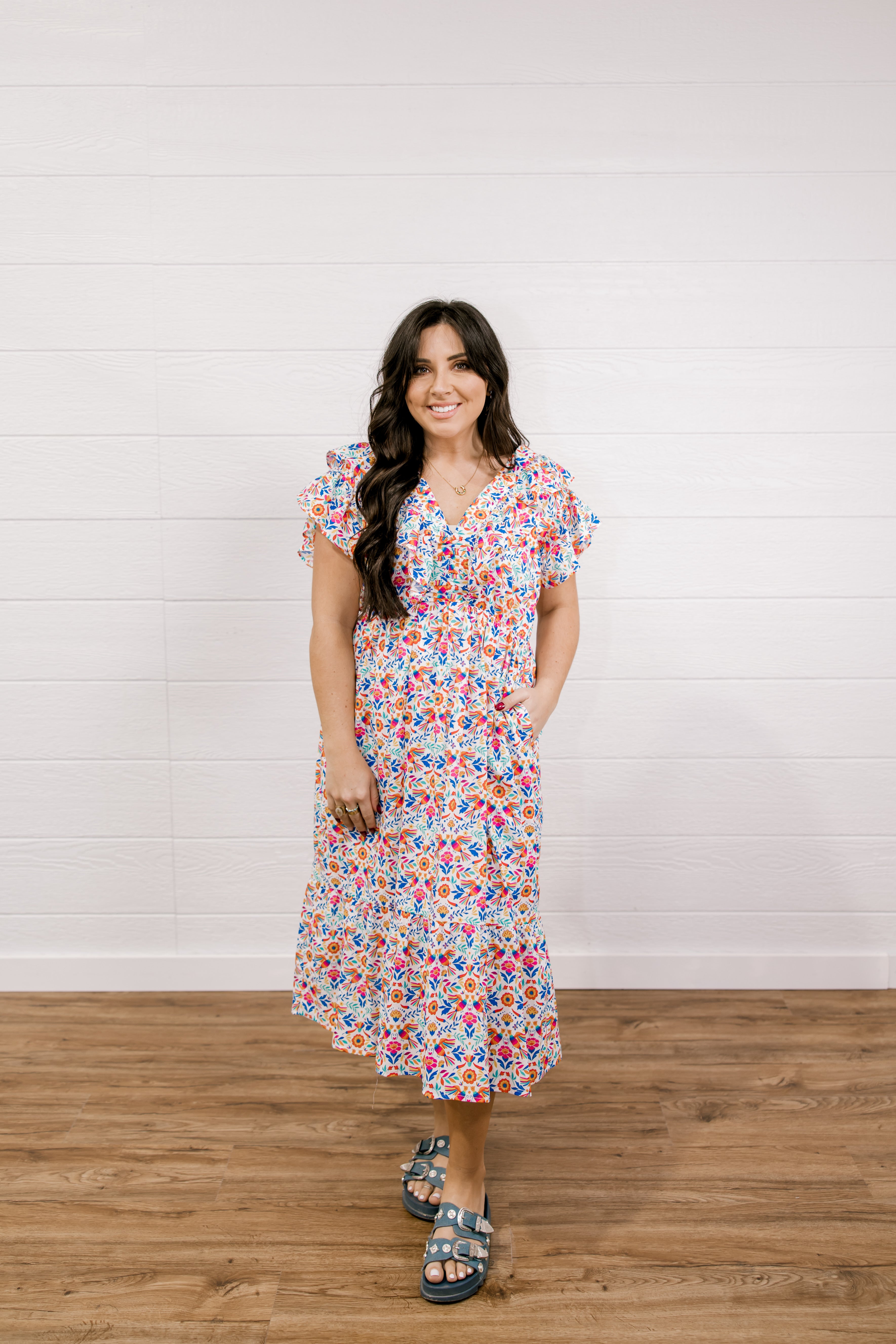 Woman wearing a colorful floral dress standing against a white wall.