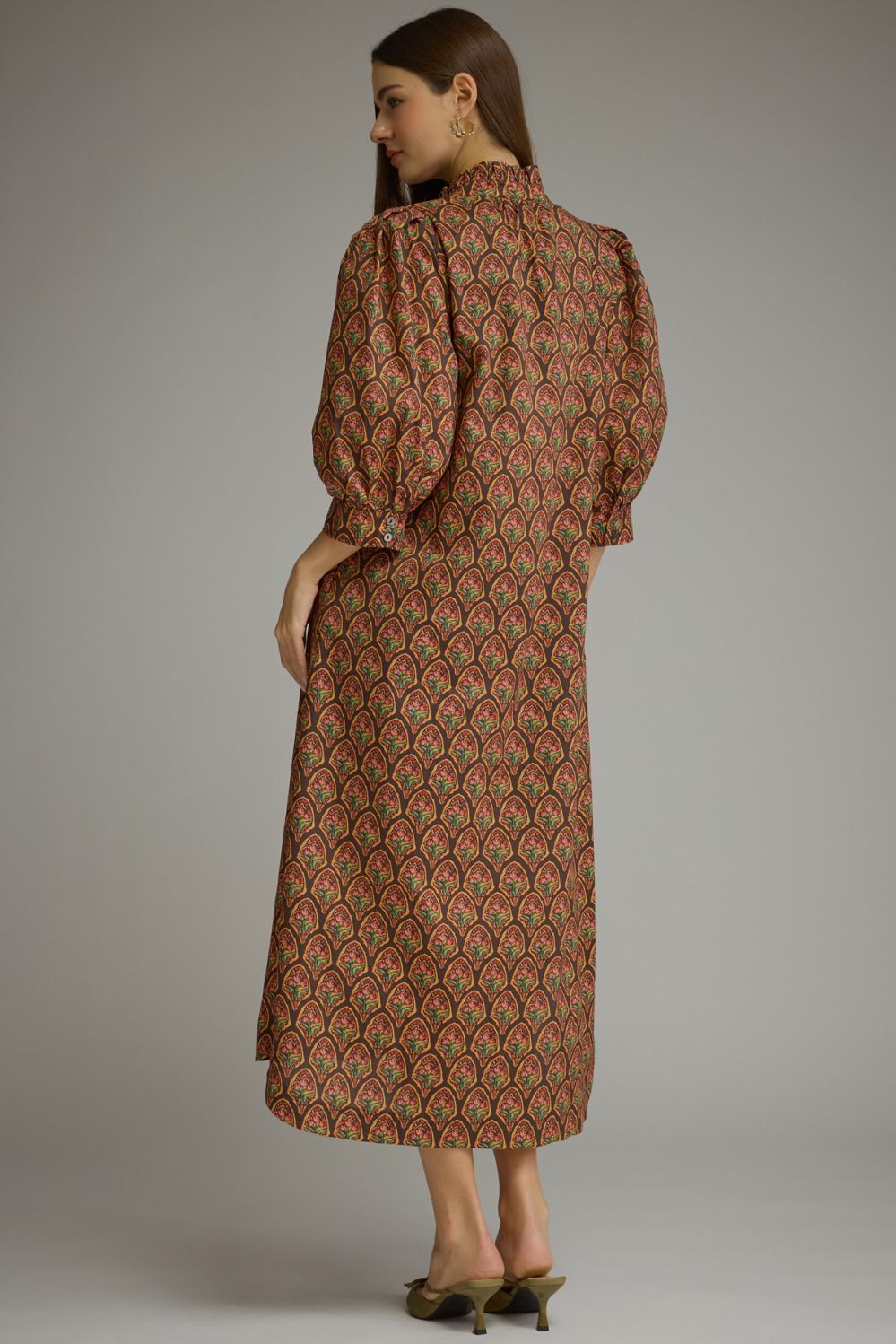 A woman modeling a long-sleeved midi dress with a brown, red, and green scalloped pattern, shown from a side-back angle against a neutral gray studio background.