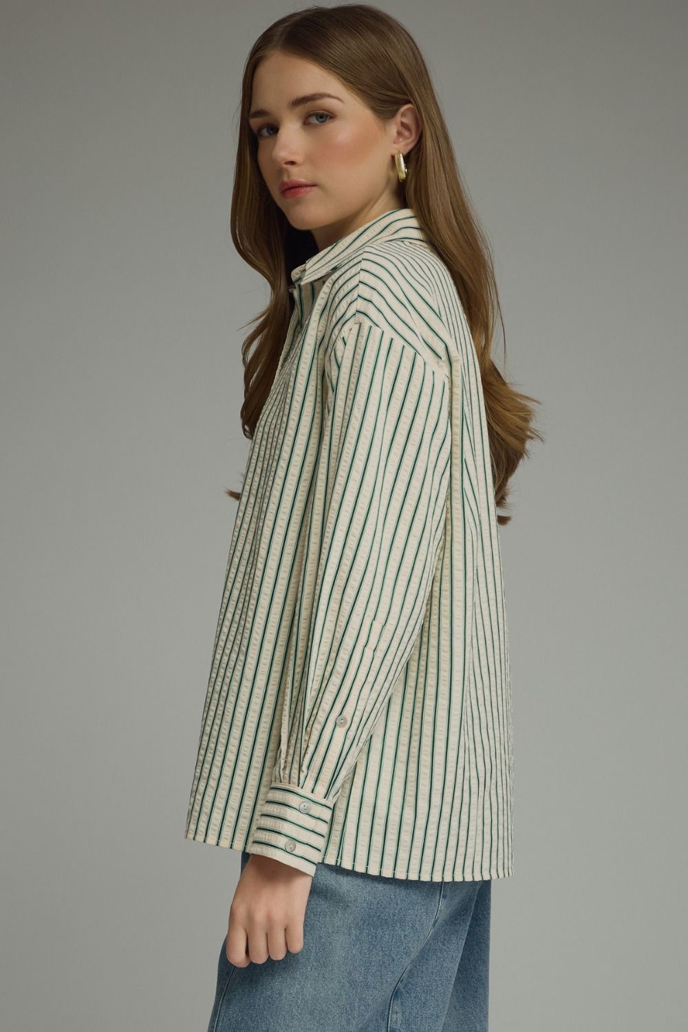 A woman wearing a long-sleeve button-up shirt with thin vertical dark green stripes on an off-white base, paired with blue jeans, photographed against a solid gray studio background.