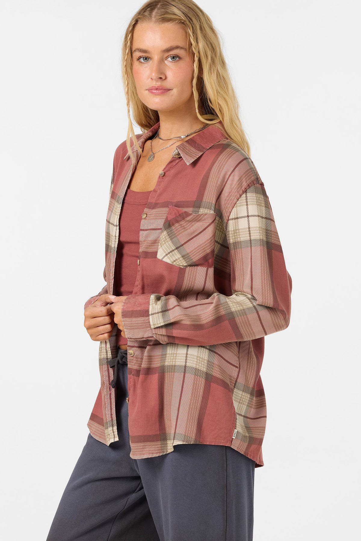 A woman modeling a long-sleeve plaid button-up shirt in dusty rose and beige tones, worn open over a matching tank top and dark gray pants, against a plain light gray studio background.