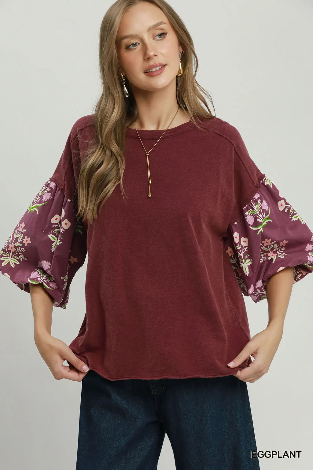 A woman wearing a burgundy top with floral bell sleeves and dark blue jeans, standing against a light gray studio background. The brand name 'EGGPLANT' is visible in the bottom right corner.