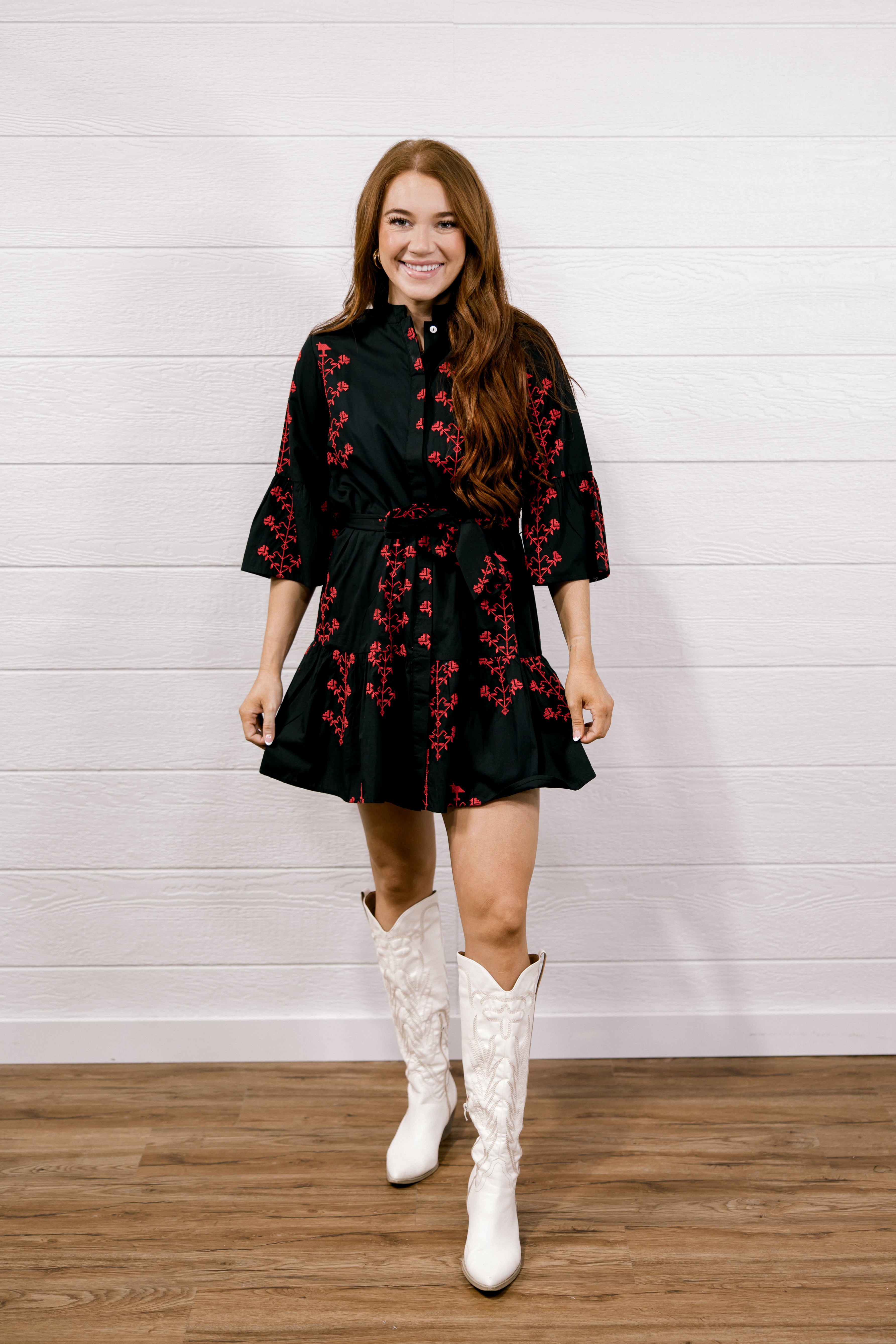 A woman modeling a black mini dress with red floral embroidery and white lace cowboy boots, standing against a white shiplap wall and wood floor.