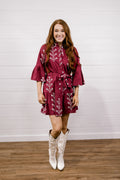 A woman modeling a short burgundy wrap dress with white floral embroidery and white cowboy boots, standing against a white wood-paneled wall and wood floor.