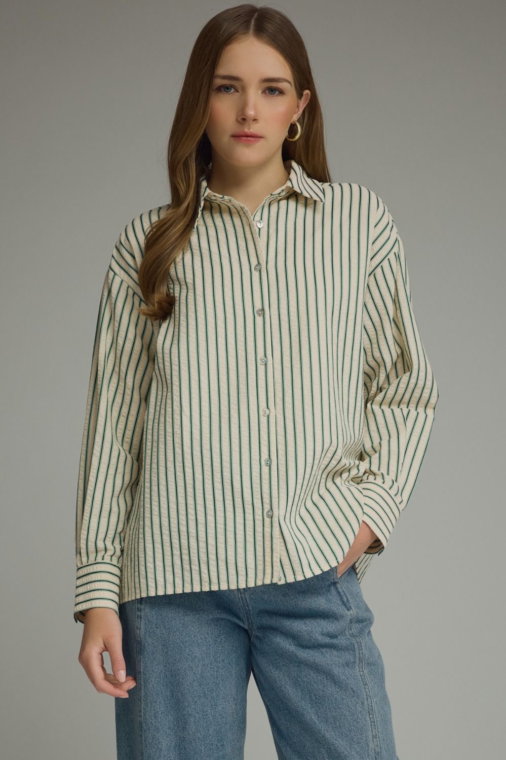 A woman wearing a long-sleeve button-up shirt with vertical stripes in green and cream, paired with blue jeans, photographed against a solid gray studio background.