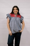 A woman modeling a short-sleeve blouse with a gray and white vertical stripe body, a red and white floral yoke, and ruffled sleeves, paired with dark jeans against a white wood-paneled wall.