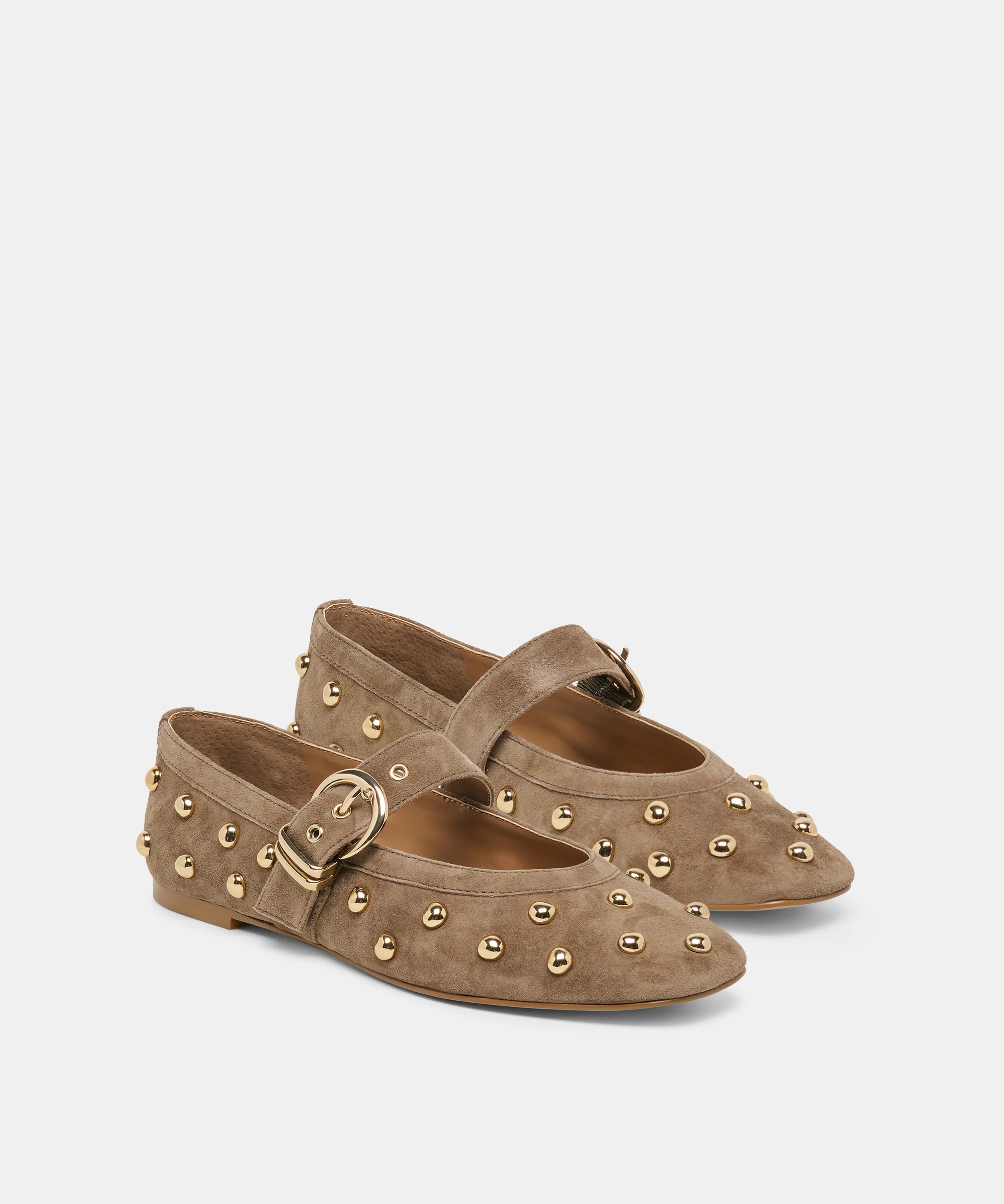 A pair of taupe suede Mary Jane flats decorated with gold-tone metal studs and buckles, shown against a light gray background.