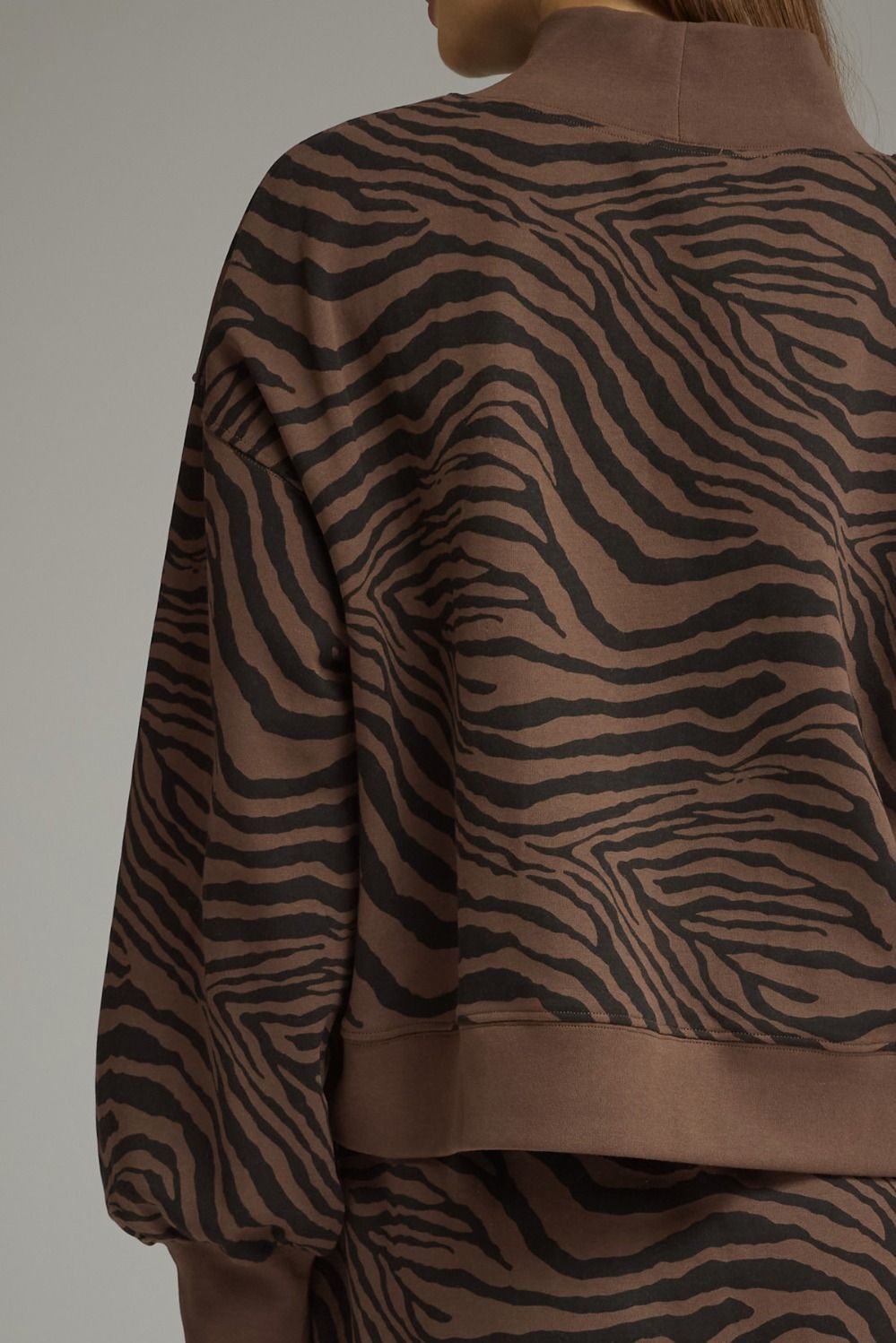 A close-up back view of a person wearing a brown and black zebra-print cropped sweatshirt with ribbed trim, against a plain light gray studio background.