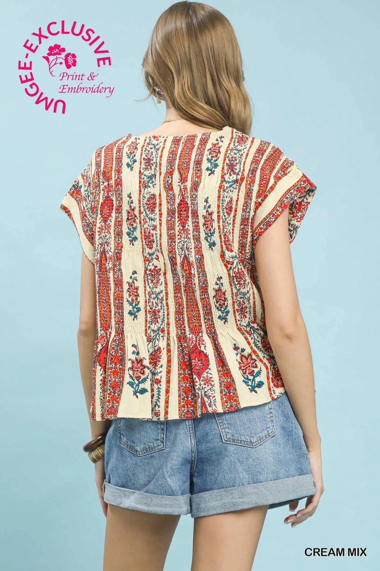 Woman wearing a cream blouse with red floral patterns and denim shorts on a light blue background.
