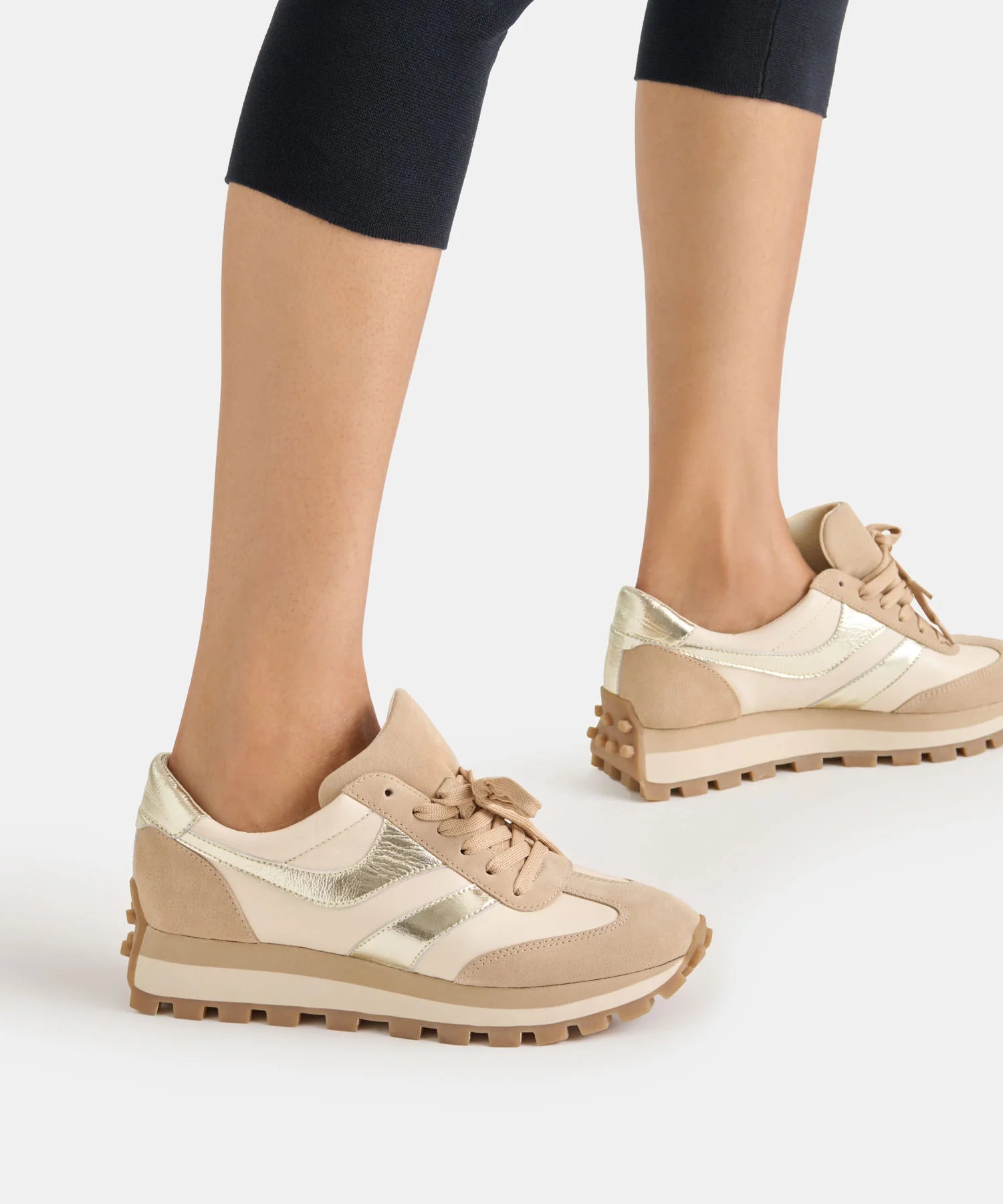 Beige and metallic gold low-top sneakers worn with black cropped leggings, shown from a side angle against a light gray studio background.