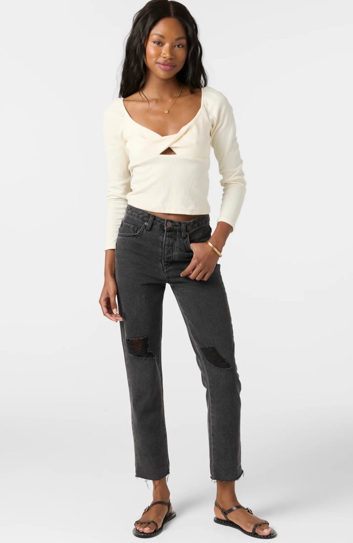 A woman modeling a cream long-sleeve knit top with a crossover front and keyhole cutout, paired with dark gray distressed jeans and black sandals, against a light gray studio background.