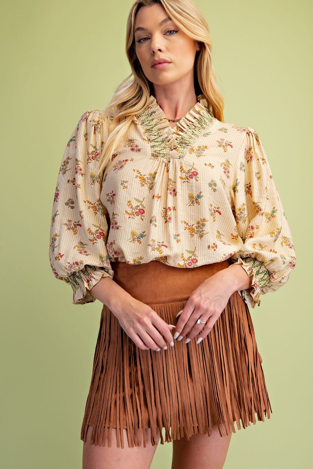 A woman modeling a cream-colored floral long-sleeve blouse with a ruffled high neck and brown fringe mini skirt, set against a solid light green background.