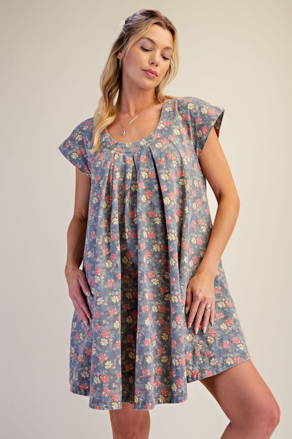 A woman modeling a short-sleeve, knee-length shift dress with a blue-gray base and a dense multicolor floral print, set against a plain beige studio background.