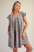 A woman modeling a short-sleeve, knee-length shift dress with a blue-gray base and a dense multicolor floral print, set against a plain beige studio background.