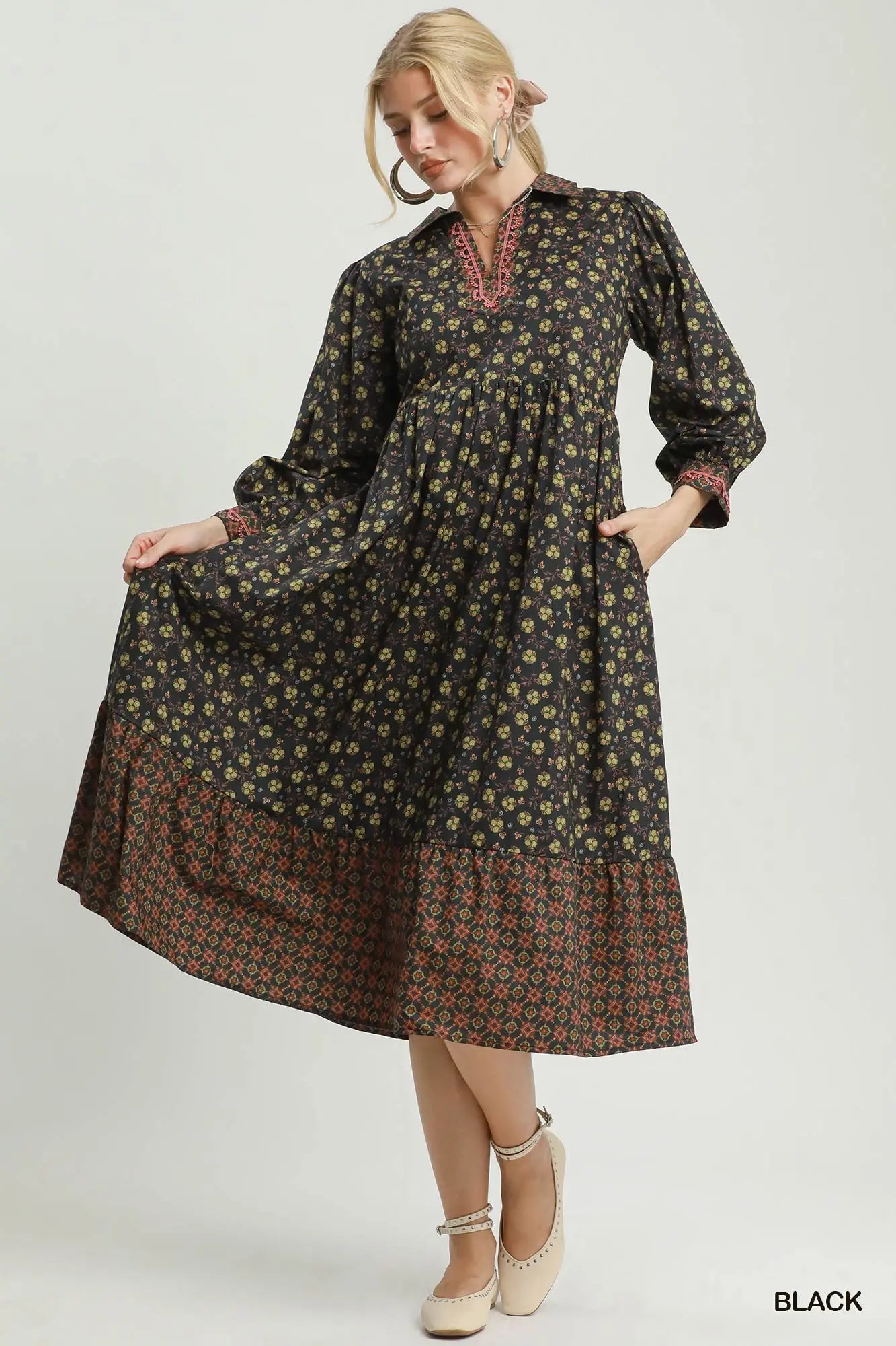 A woman modeling a long-sleeved black midi dress with a small yellow and green floral print and a contrasting brown patterned hem, set against a plain light gray background.
