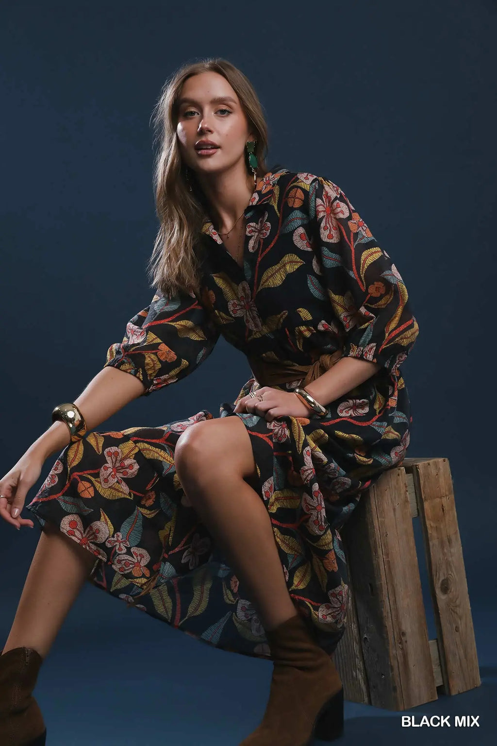 A woman models a black long-sleeve button-up dress with a vibrant multicolor floral and botanical print, sitting on a rustic wooden crate against a dark blue studio background.