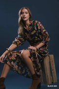 A woman models a black long-sleeve button-up dress with a vibrant multicolor floral and botanical print, sitting on a rustic wooden crate against a dark blue studio background.