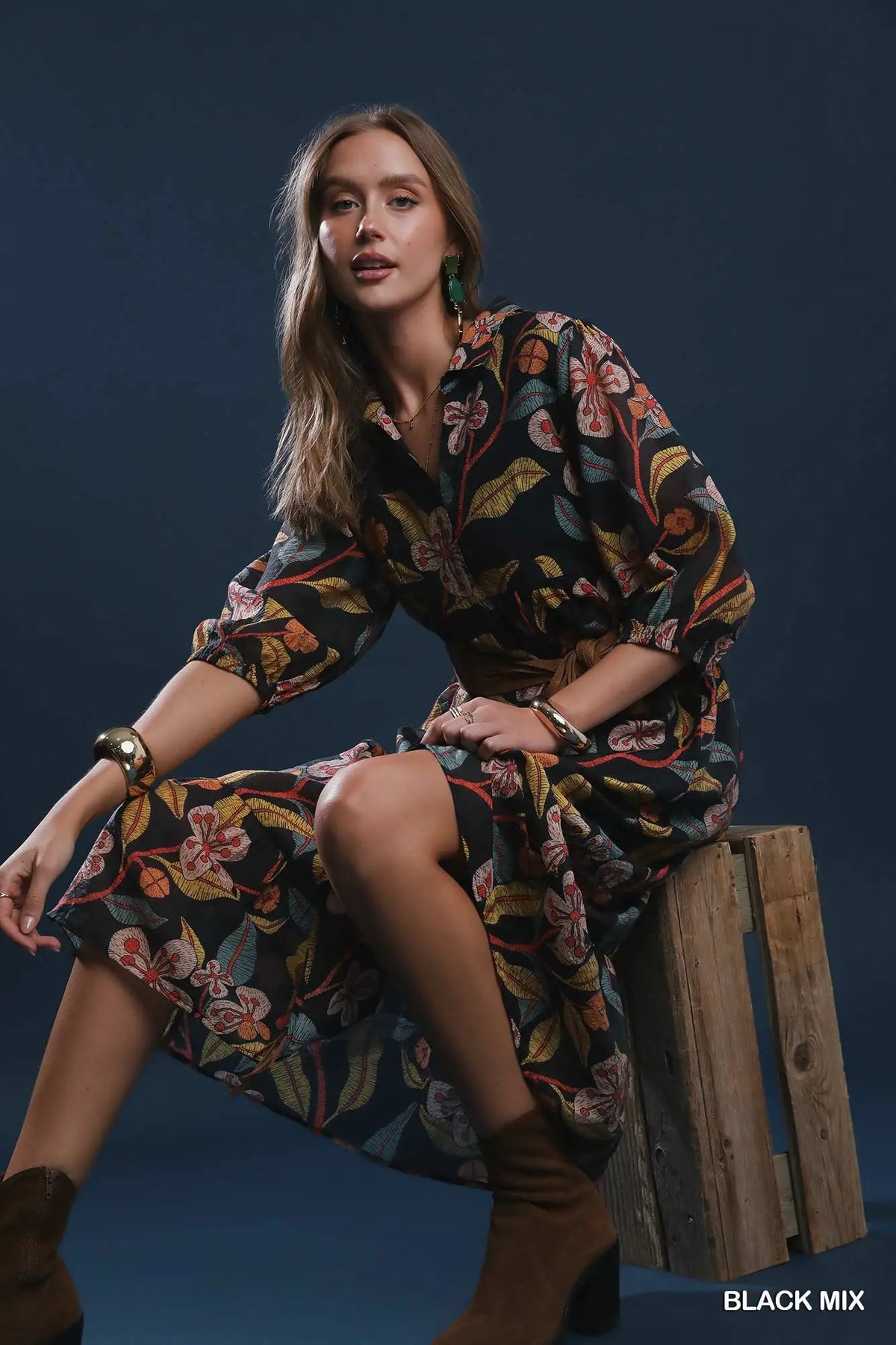 A woman models a black long-sleeve button-up dress with a vibrant multicolor floral and botanical print, sitting on a rustic wooden crate against a dark blue studio background.
